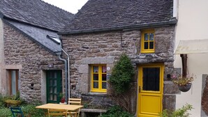 Un bâtiment en pierre avec une porte et des fenêtres jaunes, une petite table d’extérieur avec des chaises et un pot de fleurs.