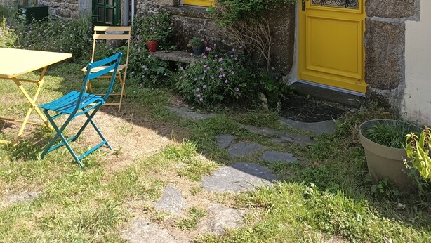 Un bâtiment en pierre avec une porte et des fenêtres jaunes, un petit jardin avec des fleurs, une chaise et une table.