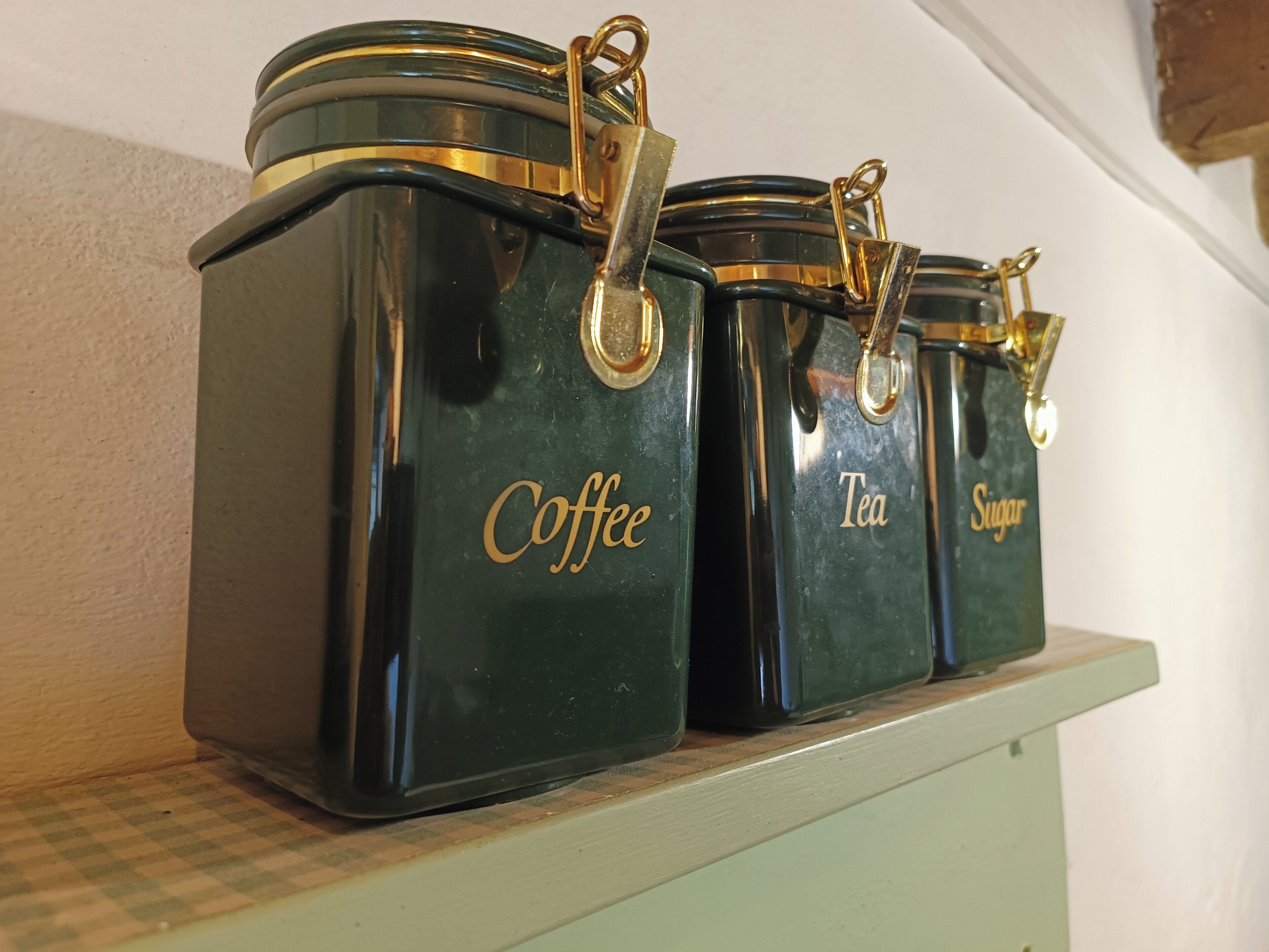 Trois récipients vert foncé, ornés de fermoirs dorés, étiquetés respectivement « Café », « Thé » et « Sucre », sont posés sur une étagère.