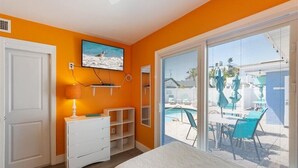 Un dormitorio con una pared naranja, una cama, un tocador blanco, un televisor y un gran ventanal corredera que da a una zona de piscina.