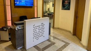 Uma área de recepção com televisão exibindo uma vista urbana, um balcão de atendimento e uma grande obra de arte abstrata na parede.
