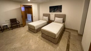 Quarto de hotel com duas camas de solteiro, uma pequena mesa, um banco e um quadro na parede.