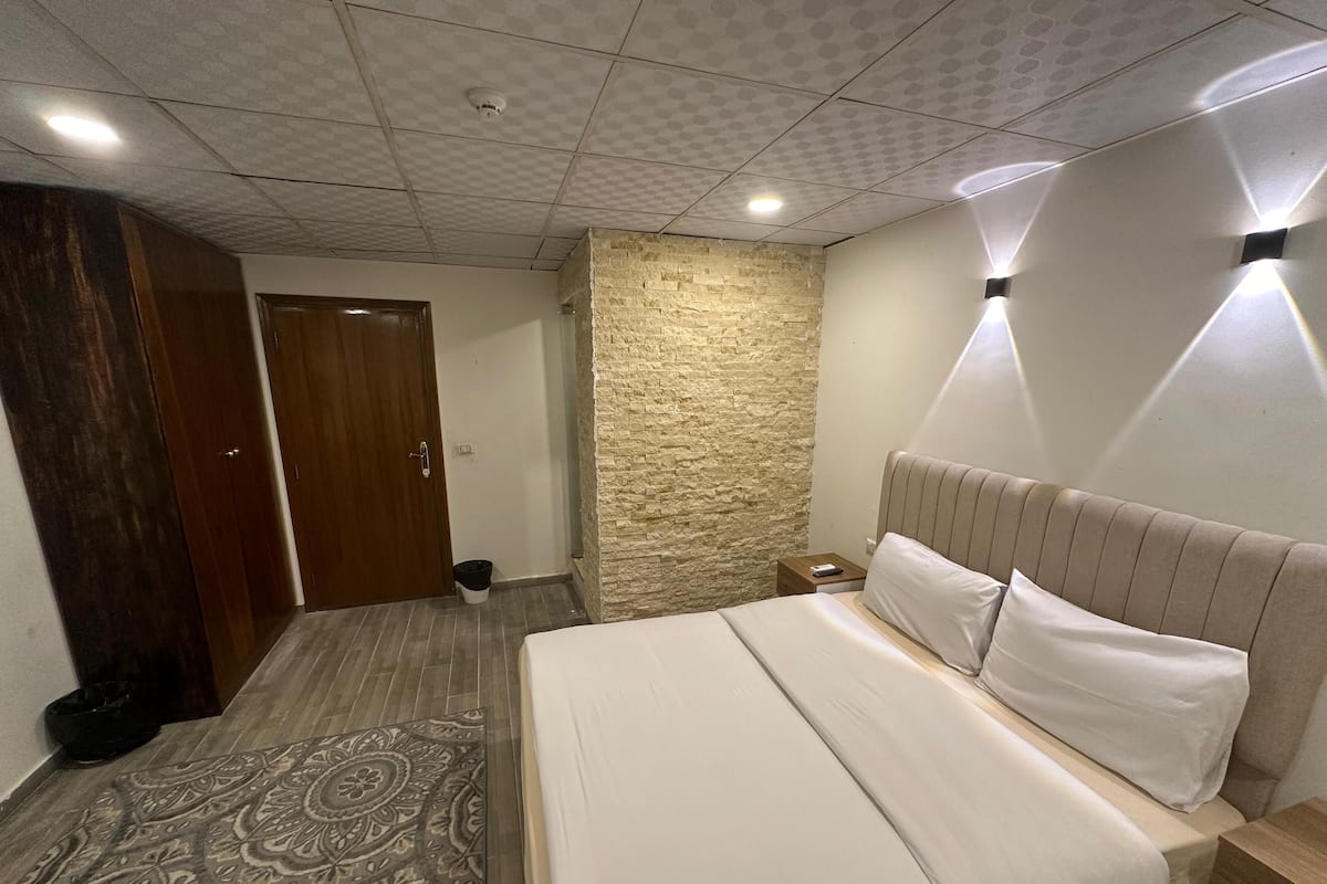 Un dormitorio con cama, mesita de noche, armario de madera, alfombra estampada, pared de piedra decorativa e iluminación empotrada en el techo.