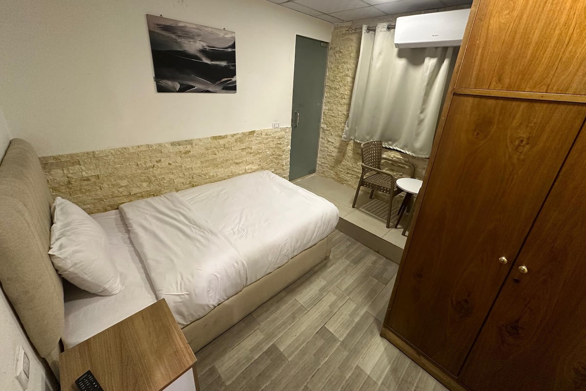 Un dormitorio con cama, armario de madera, una pequeña mesa con silla y aire acondicionado instalado en la pared.