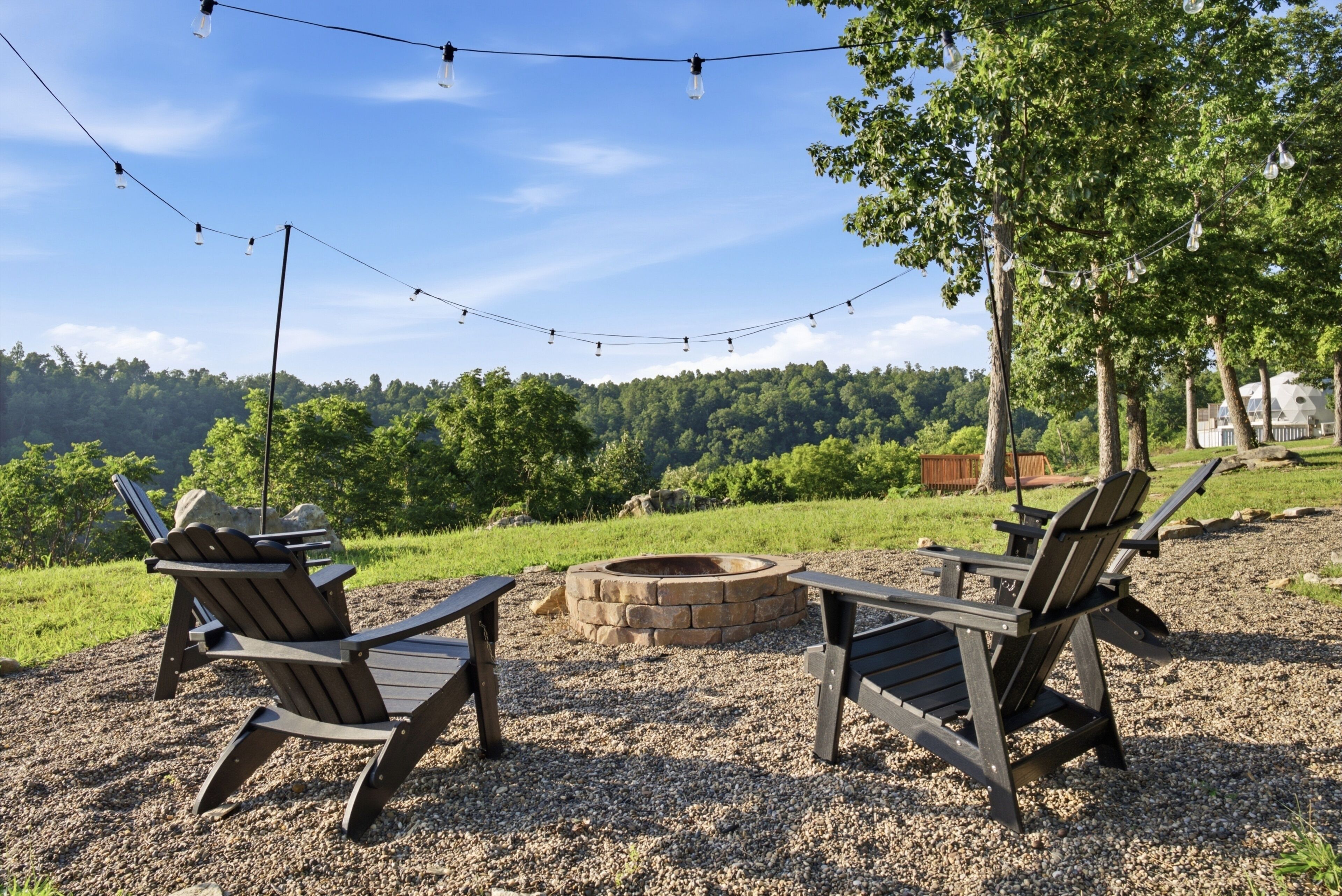 An outdoor seating area with Adirondack chairs and a fire pit, surrounded by a gravel ground and a view of a forested hill.