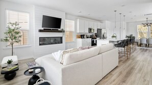 A modern living room with a large sofa, a flat-screen TV mounted above a fireplace, and a kitchen area with white cabinets and stainless steel appliances.
