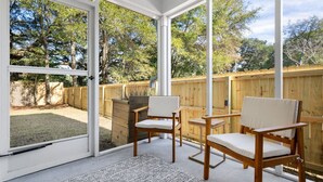 A covered patio with two wooden chairs and a small table, a wooden fence, and a view of a backyard with trees.