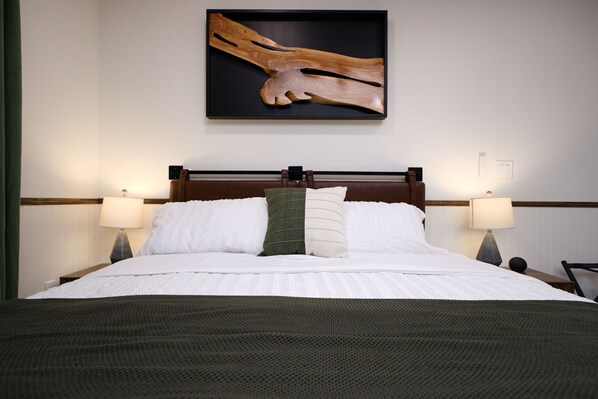 A neatly made bed with white linens and a dark green throw blanket, flanked by two bedside lamps and a wooden headboard.