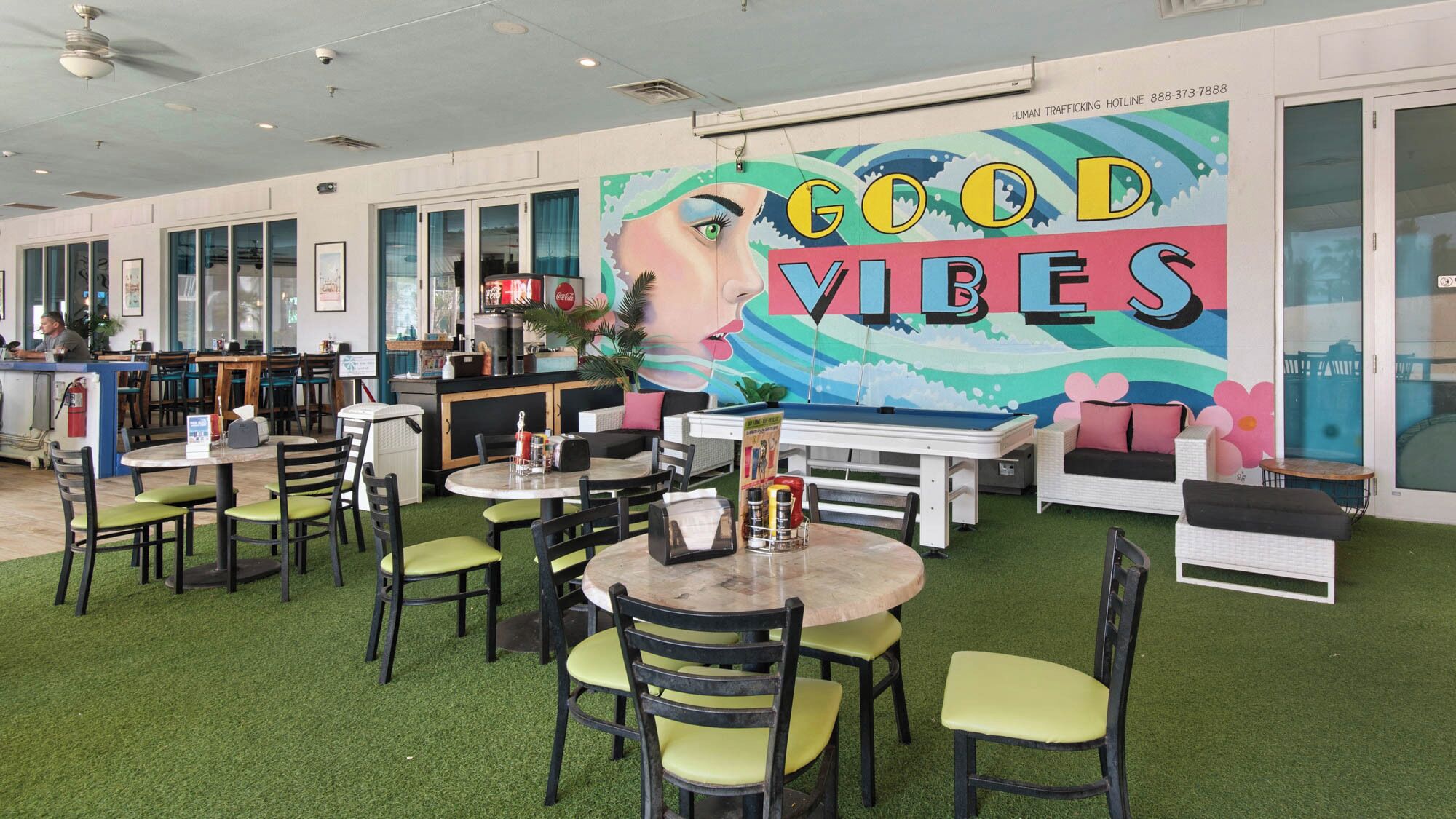Un restaurante con mesas en el interior, un mural con el texto 'GOOD VIBES' y un refrigerador de Coca-Cola.