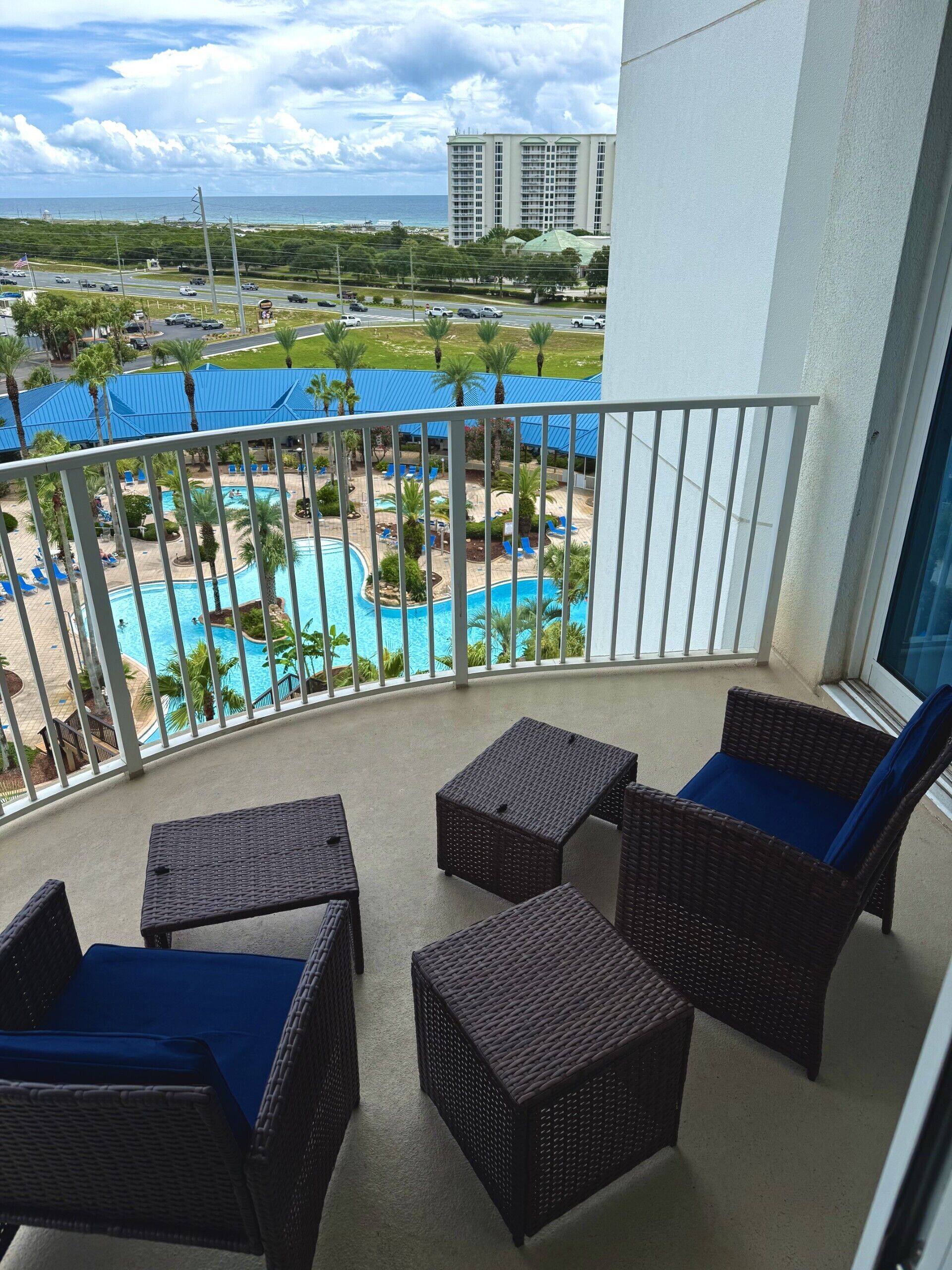 A balcony with wicker furniture and a view of a pool area with palm trees and a building in the background.