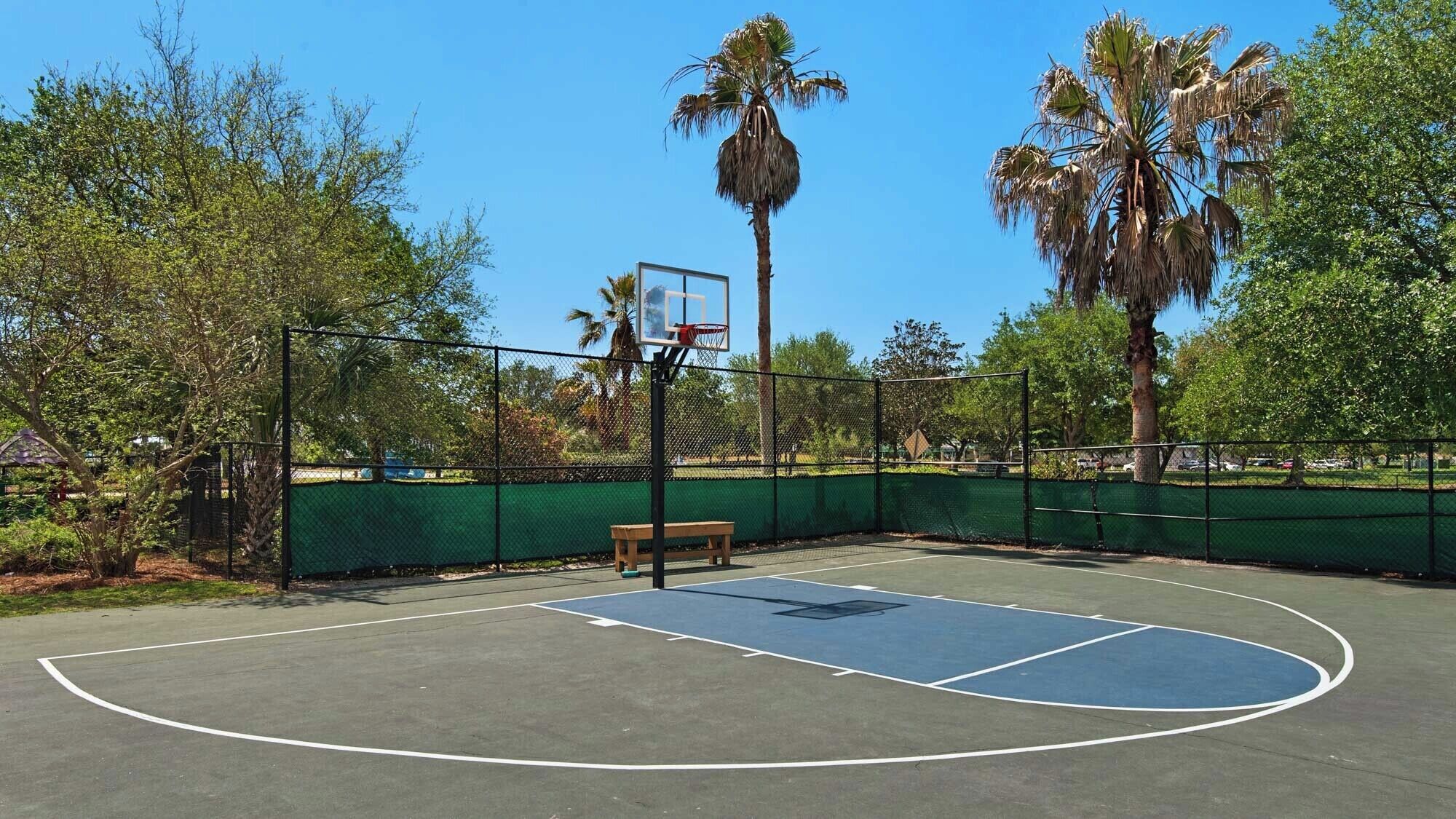 Un campo da basket all'aperto con recinzione verde, una panchina di legno e palme.