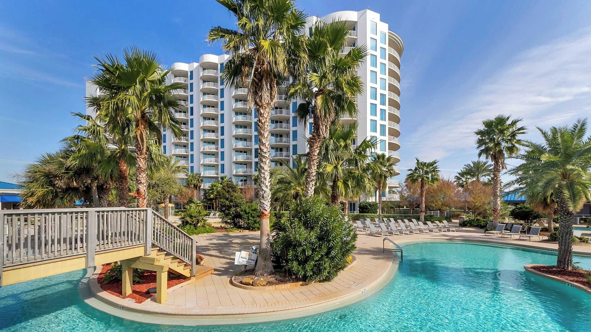 A hotel with a pool, palm trees, and a clear blue sky.