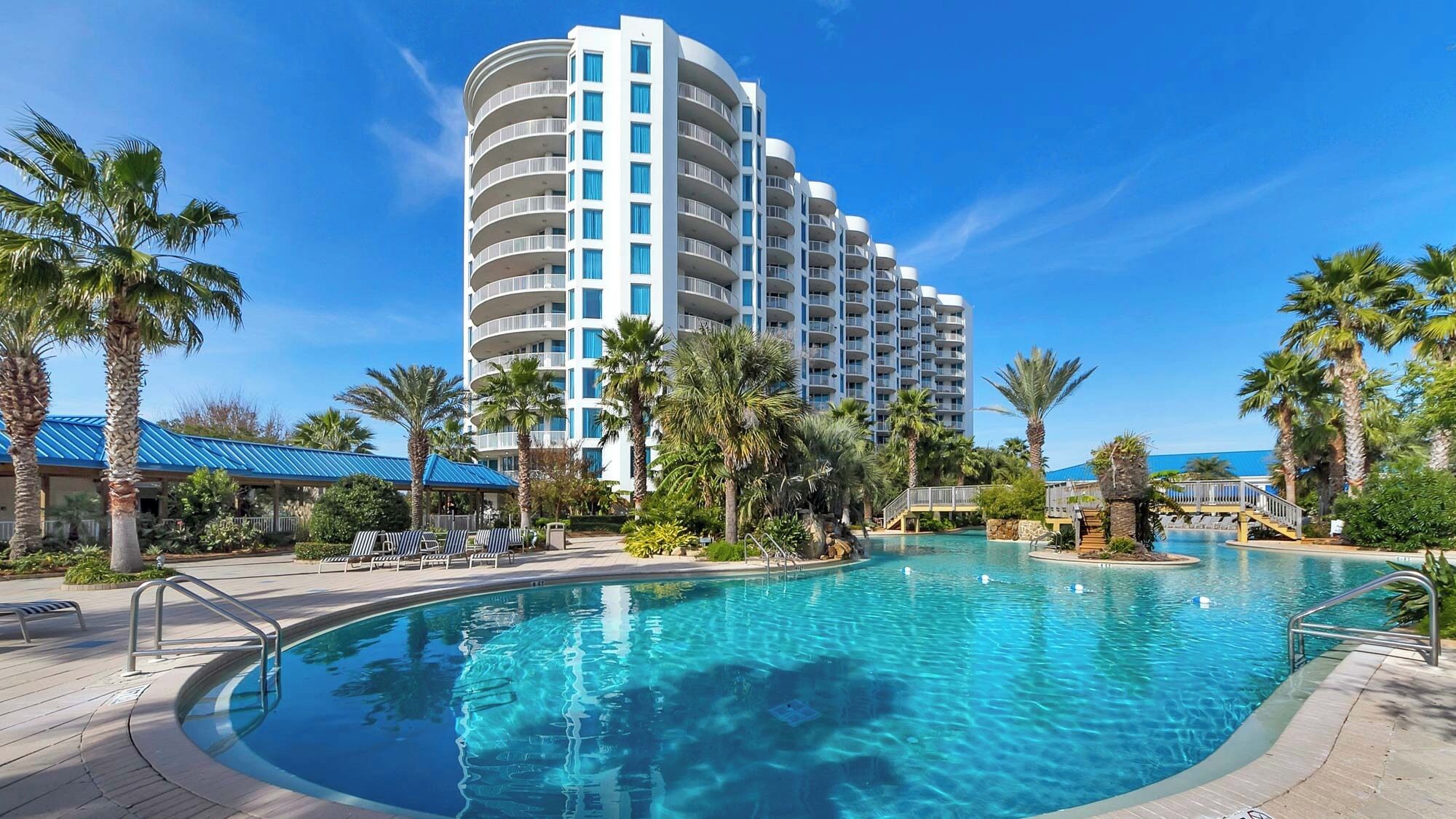 A hotel with a large pool, palm trees, and a clear blue sky.