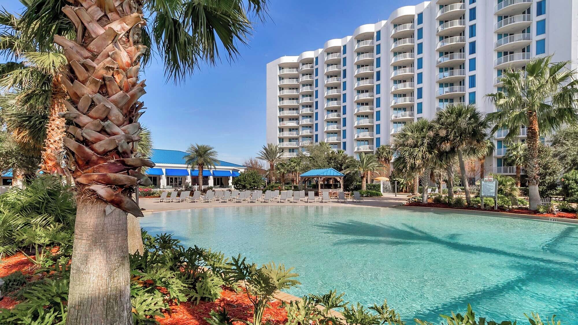 A resort with a large pool, palm trees, and a multi-story building with balconies.