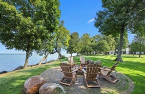 A circular outdoor seating area with Adirondack chairs and a fire pit, surrounded by trees and a view of the water. - Charming Waterfront Lake Villa on Mille Lacs Lake (Onamia)
