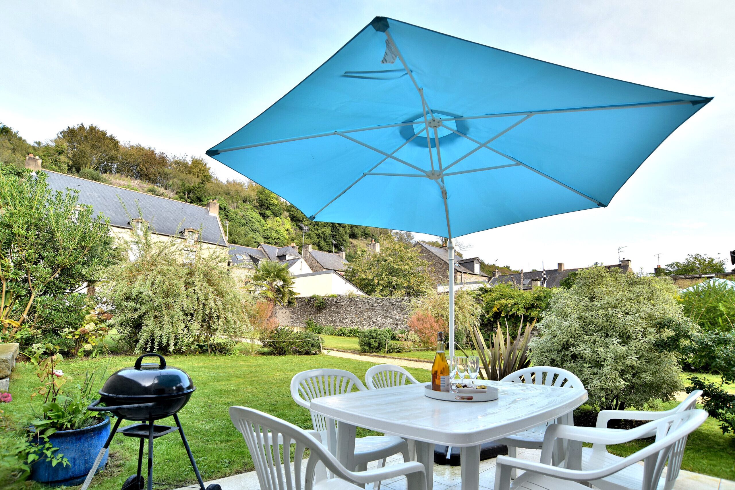 Eine Terrasse mit Tisch und Stühlen unter einem blauen Sonnenschirm, ein Grill und eine Weinflasche.