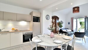 A modern kitchen with white cabinets, a microwave, and a sink. There is a dining area with a round table set for four, white chairs, and a vase with flowers. A clock is on the wall, and a wooden cabinet is visible. - La Petite Jeanne (Cancale)