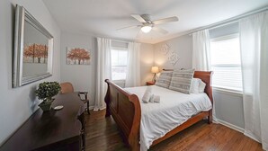 Une chambre à coucher comprenant un lit, un bureau, une chaise, un ventilateur de plafond et une fenêtre avec des rideaux.