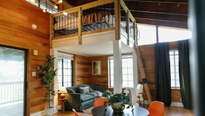 Un vaste espace de vie de style loft, doté d’un plafond haut, de murs en bois, d’un balcon, d’un coin repas avec des chaises orange et d’un escalier menant à un niveau supérieur.