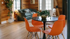 Une pièce spacieuse de style loft, dotée d’un plafond haut, d’un escalier en bois, d’un coin repas avec une table ronde et des chaises orange, d’un ensemble de canapés et d’une grande fenêtre laissant entrer la lumière naturelle.