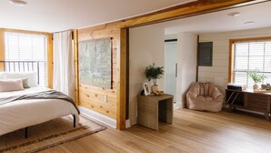 Une chambre à coucher avec un lit, une tête de lit en bois, une grande fenêtre avec des stores, un tableau, une petite table d’appoint avec une plante et un fauteuil en forme de poire.