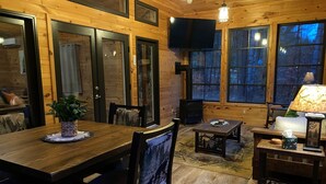 A cozy cabin interior with a wooden dining table, chairs, a fireplace, and large windows. - Pine View Retreat | Walk to Lake | Game Room (Brantingham)