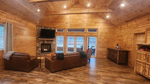 A log cabin interior with a stone fireplace, wooden walls, and a large window overlooking a deck. - Pine View Retreat | Walk to Lake | Game Room (Brantingham)