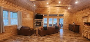 A log cabin interior with a stone fireplace, wooden walls, and a large window overlooking a deck. - Pine View Retreat | Walk to Lake | Game Room (Brantingham)