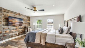 A modern bedroom with a wooden floor, a large bed, a mounted TV, and a ceiling fan. - Coastal Village Townhome | Blocks2 Grandview Beach (Encinitas)