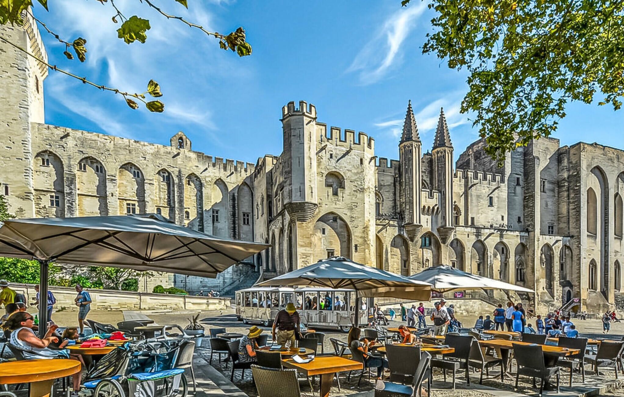 An outdoor café with tables and umbrellas in front of a historic stone building with multiple arches and towers.
