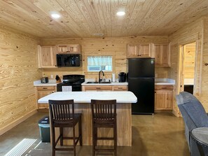 A compact kitchen with wooden cabinetry, a black refrigerator, a microwave, and a stove. There are two wooden bar stools and a small trash bin.