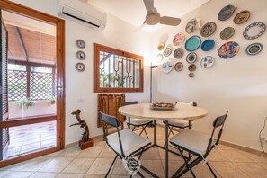 Un comedor con una mesa redonda y cuatro sillas, una pared decorativa con varios platos, una ventana con vistas a la vegetación y un ventilador de techo.