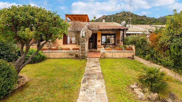 A Mediterranean-style villa with a stone pathway leading to the entrance, surrounded by lush greenery and a hilly backdrop. - Villa Mirto – Cozy Cottage with Garden and Terrace (Is Molas)