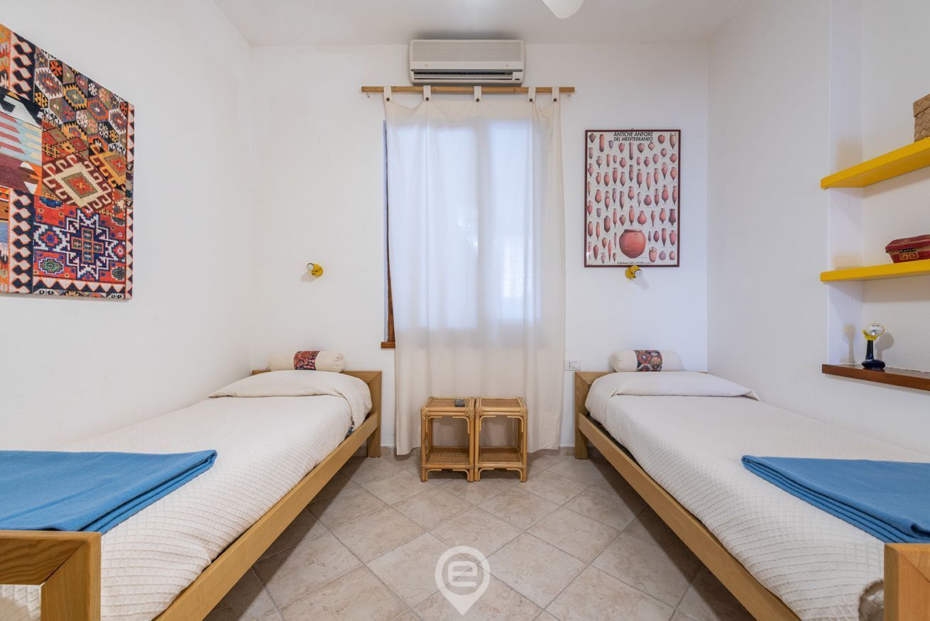 Ein Zimmer mit zwei Einzelbetten, einem kleinen Tisch, einem Vorhang sowie einer Wand mit einem farbenfrohen Wandteppich und einem gerahmten Bild.