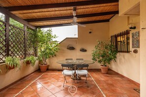 Un patio cubierto con una mesa redonda y sillas, rodeado de plantas en macetas y un muro decorativo.