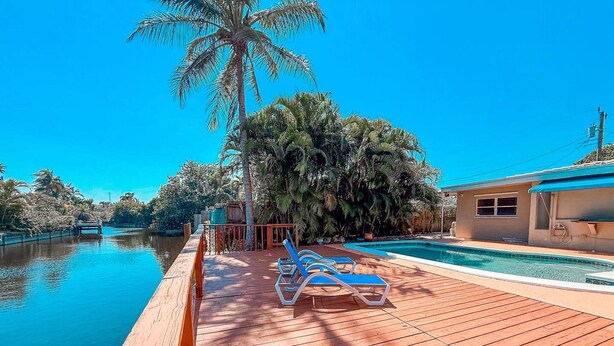 A wooden deck with a pool, palm trees, and a building with a blue roof. - Waterfront Florida Vibes and Views - King Bed (Fort Lauderdale)