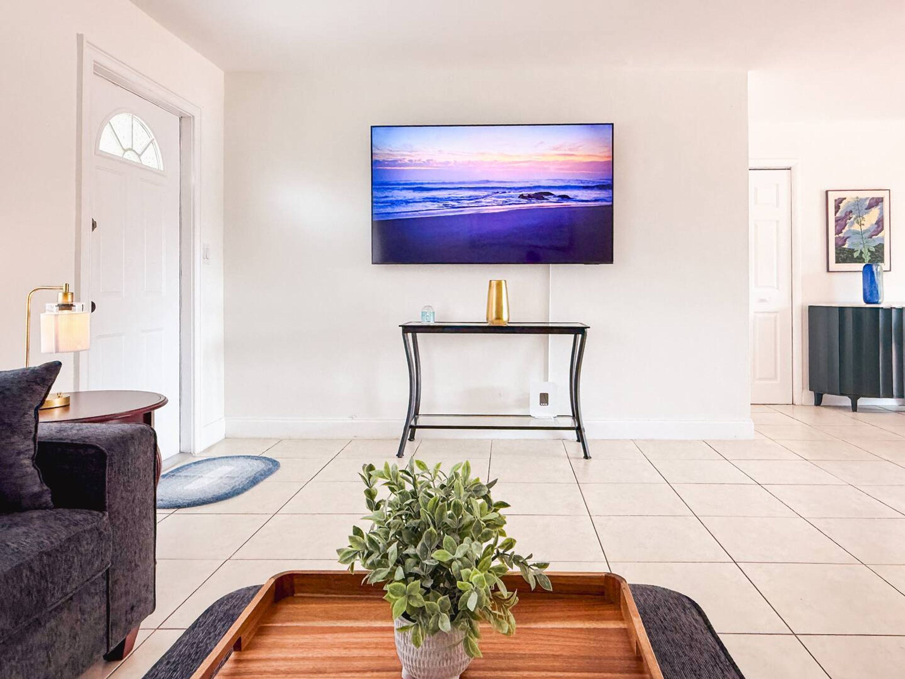 A living room with a flat-screen TV mounted on the wall, a small console table with a vase, a grey sofa, and a wooden coffee table with a plant.