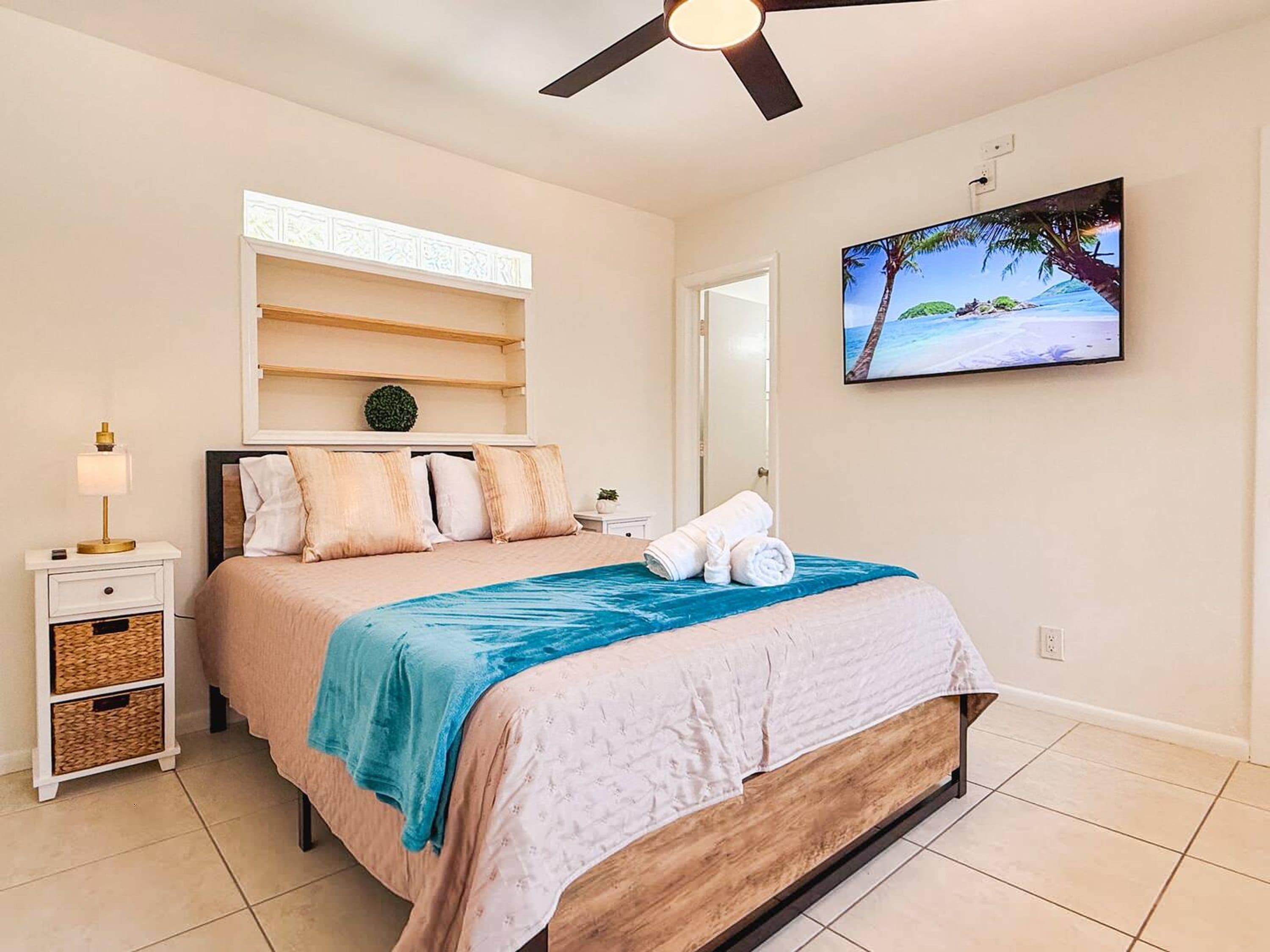 Een slaapkamer met een bed, een plafondventilator, een nachtkastje, een lamp, een aan de muur bevestigde televisie en een plank met decoratieve objecten.