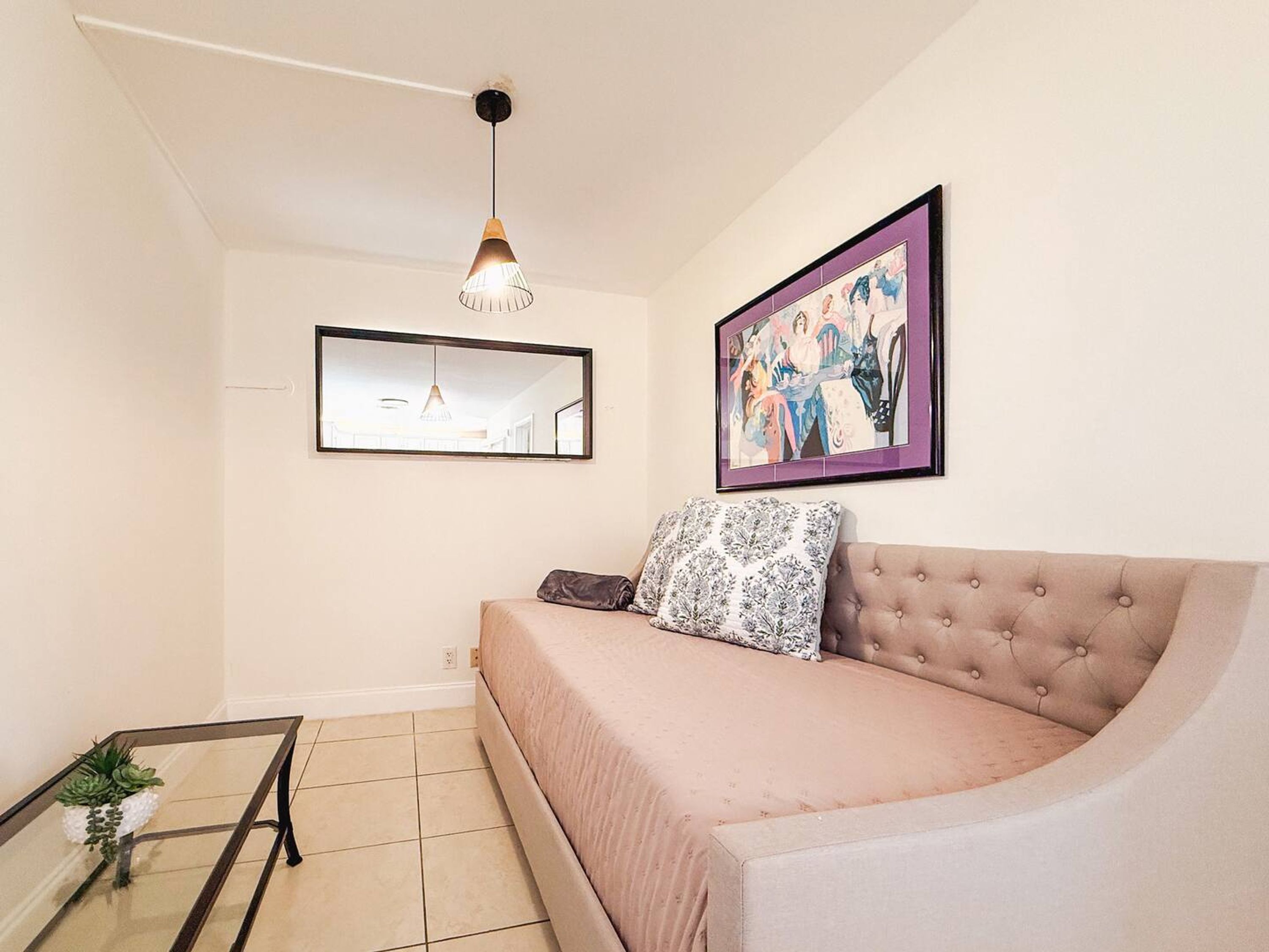 A modern living room with a beige sofa, a glass coffee table, a hanging pendant light, a framed abstract painting, and a mirror on the wall.