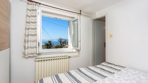 A bedroom with a bed, a window with curtains, a radiator, and a view of the sea. - Cozy apartment in Kraljevica with house sea view (Kraljevica)