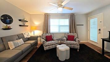 Ein Wohnzimmer mit einer Couch, zwei gemusterten Sesseln, einem Couchtisch, einem Deckenventilator, einer Uhr und einem Fenster mit Vorhängen.