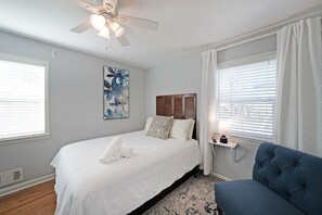 Ein Schlafzimmer mit einem Bett, einem Deckenventilator, einem Nachttisch mit Lampe und einem Fenster mit Jalousien.