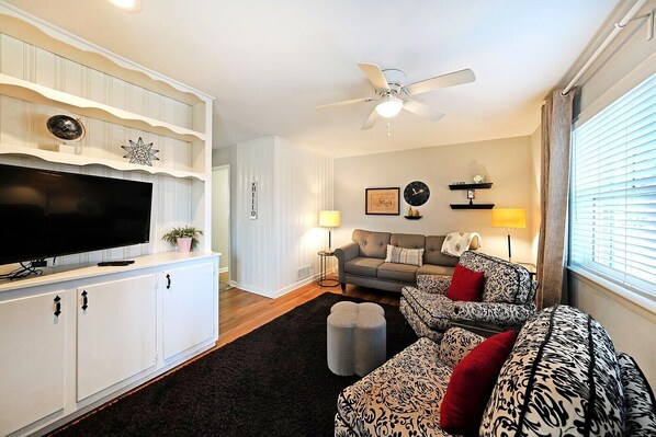 A living room with a flat-screen TV, two sofas, a ceiling fan, and a window with blinds.