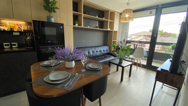 A modern kitchen with a built-in oven and a dining area with a black sofa and a dining table set for four. - Coastal apartment with lake view, heated swimming pool and jacuzzi (Puerto Varas)