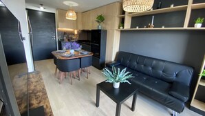 A modern living room with a black sofa, a dining table with chairs, a coffee table with a plant, and a kitchen area with cabinets and a hanging light fixture. - Coastal apartment with lake view, heated swimming pool and jacuzzi (Puerto Varas)
