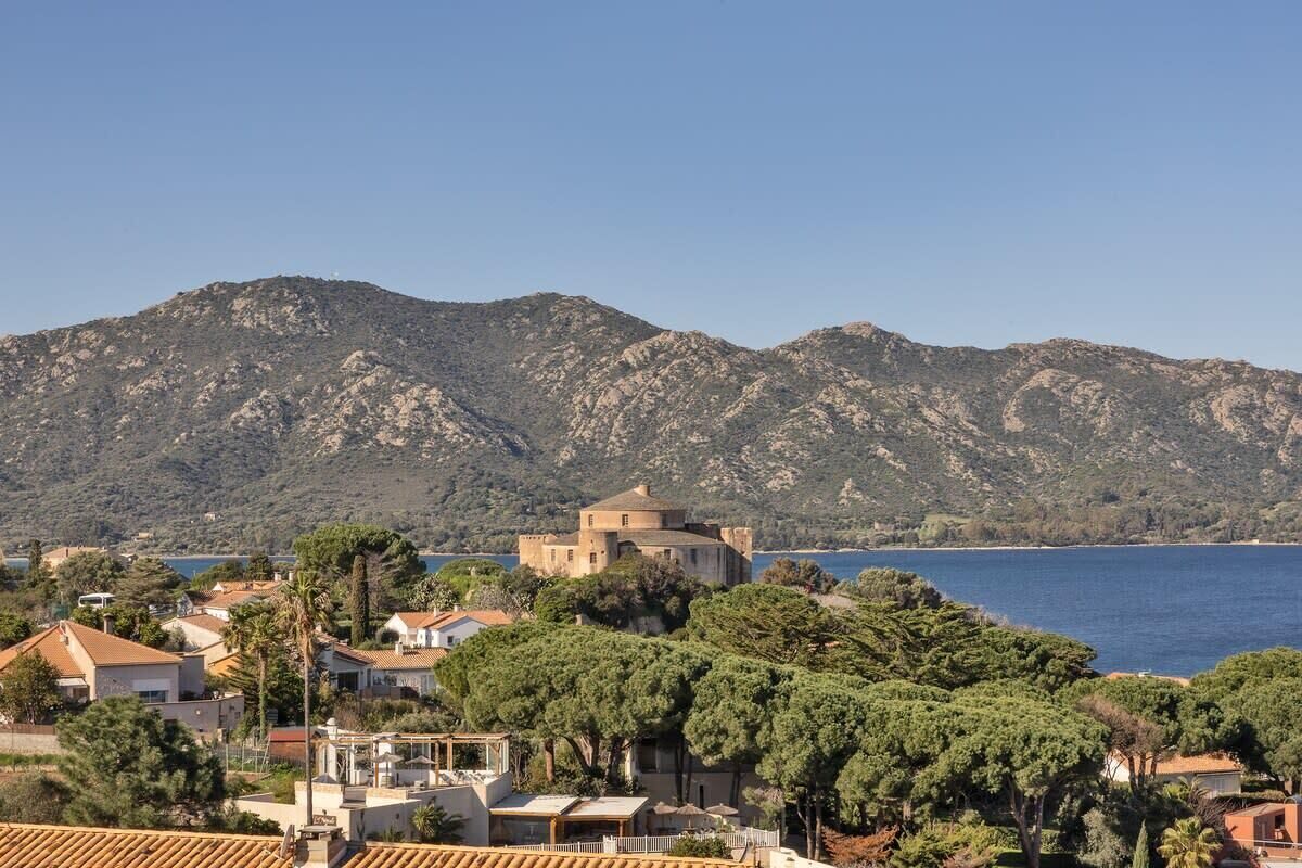 A coastal town with a prominent building surrounded by lush greenery and a backdrop of mountains.