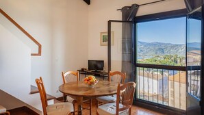 Un comedor con una mesa redonda, sillas y un frutero. Hay un televisor sobre una mesita y una ventana grande con vistas al campo.
