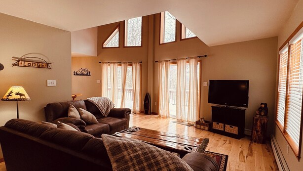 Una sala de estar acogedora con un sofá de cuero marrón, una mesita de centro y un mueble para la televisión. Hay ventanas grandes con cortinas, una chimenea y un letrero que dice "Lodge".
