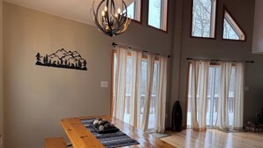 Un comedor amplio con una mesa y bancos de madera, ventanas grandes con cortinas y una lámpara de araña.