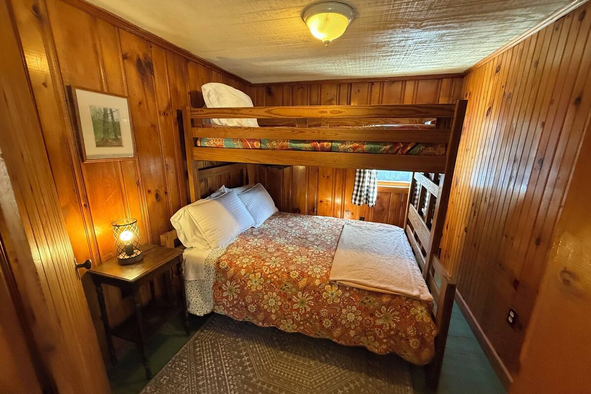A bunk bed room with wooden walls, a bedside table with a lamp, and a patterned rug.